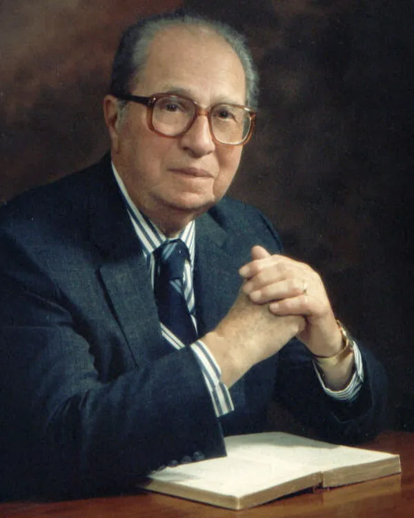 Mortimer Adler, de óculos e terno, com as mãos juntas diante de um livro aberto.
